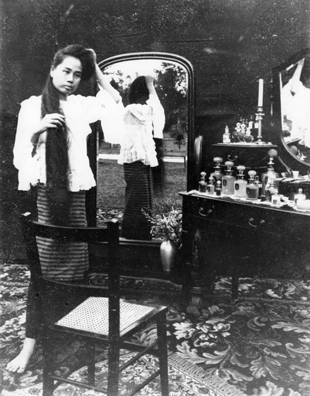 Dara Rasamee unwinding her hair before the full-length mirror. c1902.
